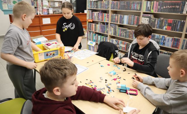 Na zdjęciu znajduje się piątka dzieci przy złączonym stole i wspólnie składają robota z klocków LEGO, które znajdują się na stole. W tle znajdują się półki z książkami.