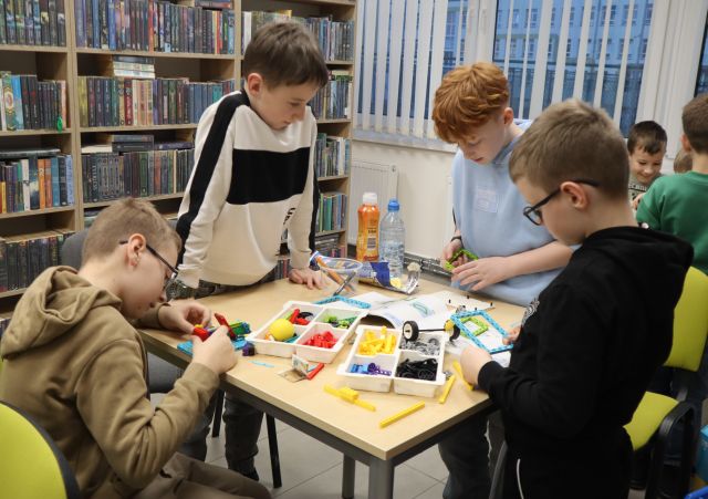 Na zdjęciu znajduje się czwórka chłopców przy stoliku, trzech z nich stoi, jeden siedzi i wspólnie układają znajdujące się na stoliku LEGO, W tle znajdują się półki z książkami oraz okna.