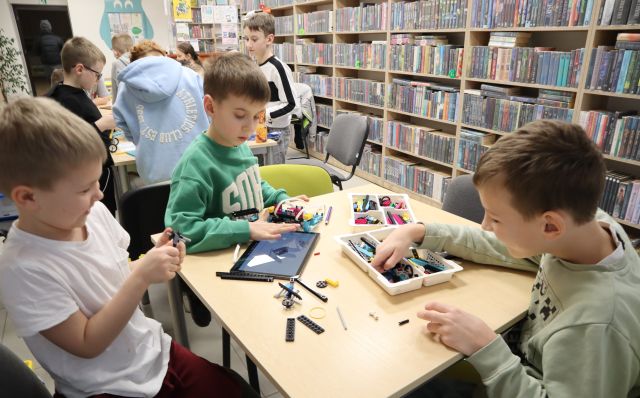 Na zdjęciu znajduje się trójka chłopców przy stoliku i wspólnie układają robota z klocków LEGO Education z klocków, które znajdują się na stole wraz z tabletem, na którym wyświetlana jest instrukcja. W tle znajdują się półki z książkami oraz inni uczestnicy zajęć.