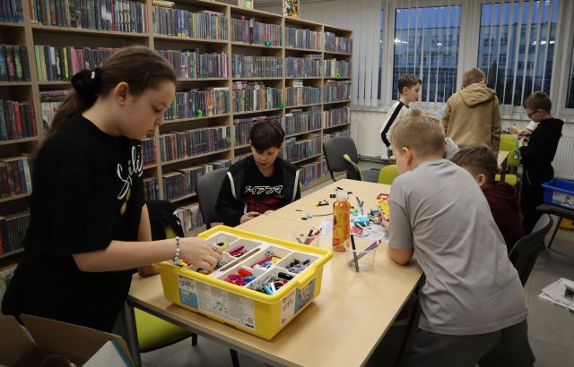 Na zdjęciu znajduje się czwórka dzieci przy złączonych stołach, dwójka z nich stoi i wszyscy wspólnie układają LEGO Education, w tle znajduje się reszta uczestników zajęć, okna a po lewej stronie znajdują się półki z książkami.