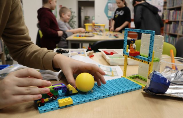 Na pierwszym planie zdjęcia znajduje się leżąca na stole konstrukcja z klocków LEGO oraz żółta piłeczka, a w tle inne dzieci uczestniczące w zajęciach.