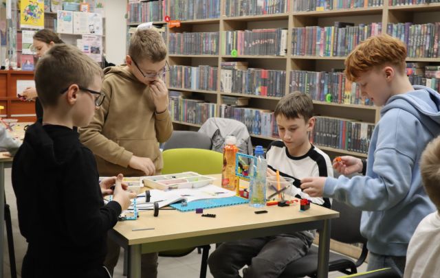 Na zdjęciu znaajduje się czwórka chłopców przy stole, na którym leżą klocki LEGO, trzech chłopców stoi, a jeden siedzi przy stole. W tle znajdują się półki z książkami.