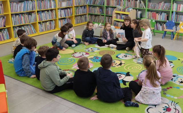 Na zdjęciu znajdują się siedzące na dywanie przedszkolaki wraz z kobietą, która trzyma w ręce książkę. W tle znajdują się półki z książkami.