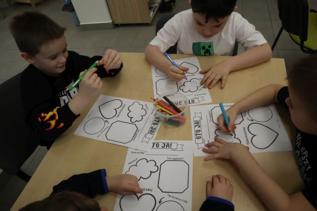 Na zdjęciu zrobionym od góry znajdują się chłopcy w wieku przedszkolnym, którzy wykonują karty pracy, na środku leżą kredki.