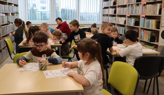 Na zdjęciu znajdują się dzieci w wieku przedszkolnym przy stolikach przy których wykonują karty pracy dotyczące emocji. W tle znajdują się okna oraz półki z ksiażkami.