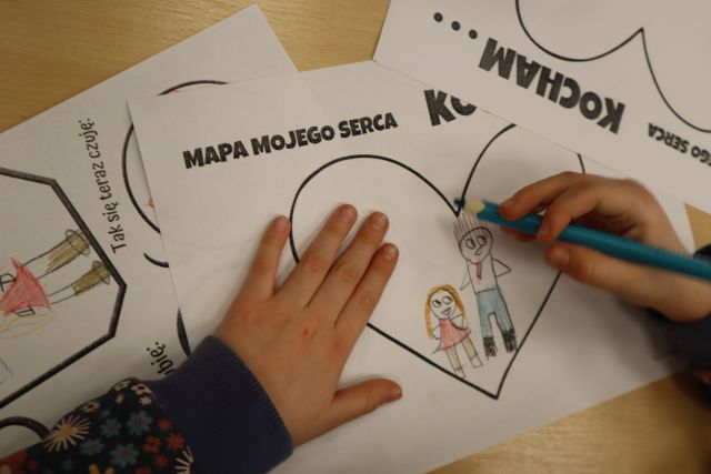 Na zdjęciu znajdują się dłonie dziewczynki, która rysuje siebie i swojego tatę w serduszku, obok niej leżą dwie prace jedna z ich przedstawia serce. Zdjęcie zostało wykonane od góry.