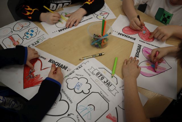 Na zdjęciu znajdują się ręce przedszkolaków, którzy malują i rysują w sercach to co kochają, obok nich leżą skończone prace dotyczące ich osoby, a na środku stołu stoją kredki.