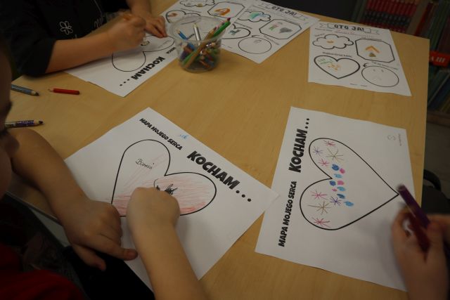 Na zdjęciu znajdują się dłonie dzieci, które malują na kartach znajdujących się na stole to co kochają w sercach. Na stoliku leży pięć prac.