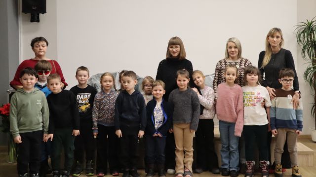 Na zdjęciu znajduje się grupa czternastu przedszkolaków wraz z czterema kobietami z tyłu. Zdjęcie zostało wykonane na holu.