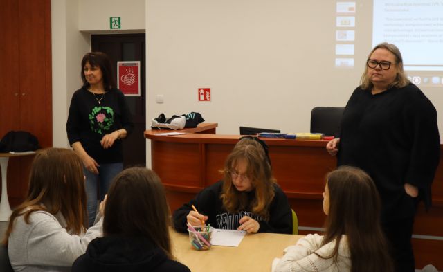 Na zdjęciu znajdują się dwie stojące przy biurku kobiety, za nimi na ścianie wyświetlana jest przentacja, a przed kobietami znajdują sie cztery dziewczyny przy stoliku.