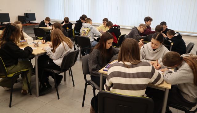 Na zdjęciu znajduje się pięć stolików, przy których siedzi młodzież i kolorują. Warsztaty odbywają się w sali komputerowej, a w tle znajdują się okna.