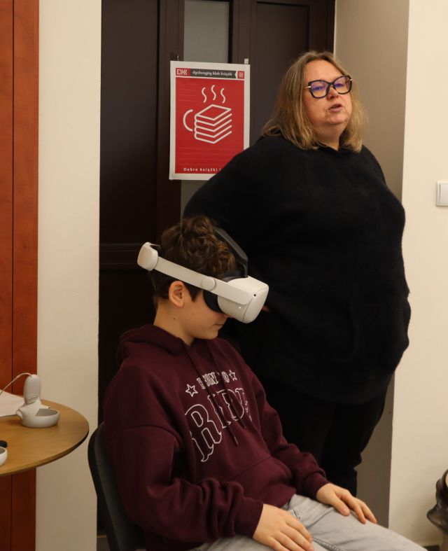 Na zdjęciu znajduje się chłopiec na krześle w okularach VR, a obok niego stoi kobieta.