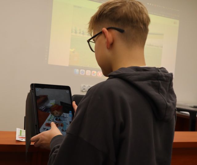 Na zdjęciu znajduje się chłopak z tabletem w ręce, którym skanuje i ożywia wykonany przez siebie rysunek. Na ścianie wyświetlana jest prezentacja. Zdjęcie zostało wykonane od tyłu.