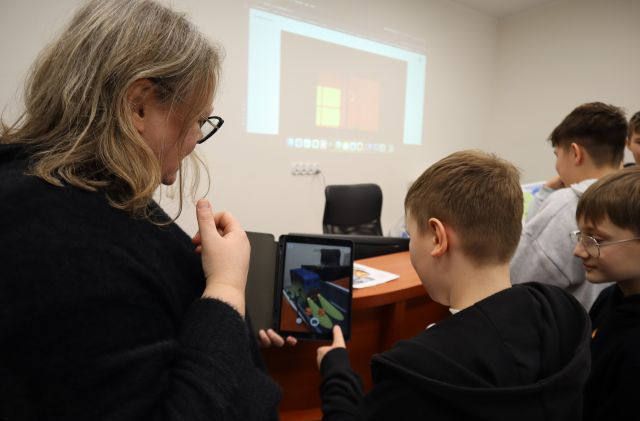 Na zdjęciu znajduje sie chłopiec, który za pomocą tabletu ożywia swój obrazek, po prawej stronie stoi w młodzież w kolejce, a po lewej stronie stoi kobieta. Na ścianie wyświetlana jest prezentacja, a zdjęcie zostało wykonane od tyłu.