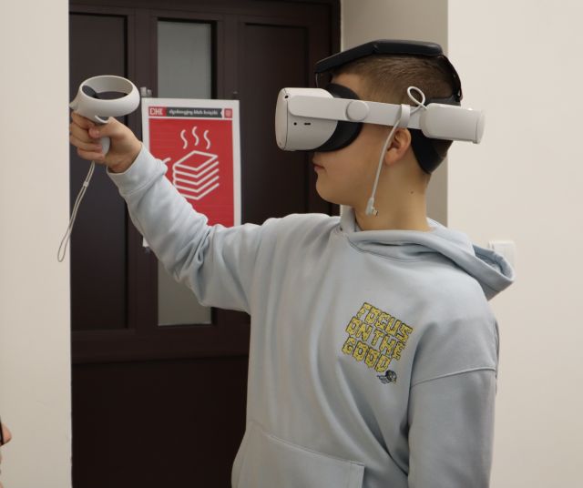 Na zdjęciu znajduje się chłopiec bokiem w okularach VR, który w uniesionej ręce na poziomie twarzy trzyma kontroler. W tle znajdują się drzwi z plakatem.