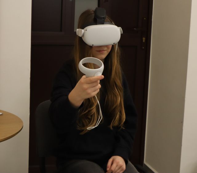 Na zdjęciu znajduje się siedząca dziewczyna w okularach VR i z kontrolerem, który trzyma w uniesionej ręce. Za dziewczyną znajdują się drzwi.
