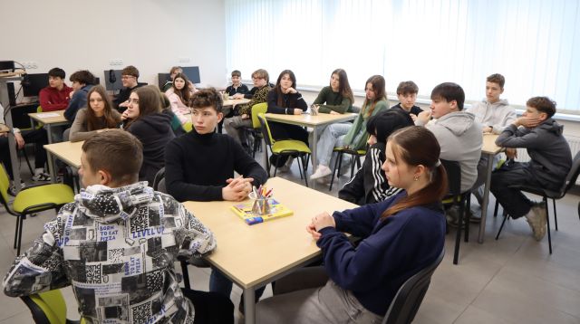 Na zdjęciu znajduje się młodzież przy stolikach, na których leżą kredki. Warsztaty odbywają się w sali komputerowej.