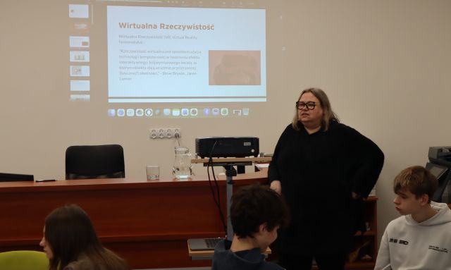 Na zdjęciu znajduje się kobieta stojąca przy rzutniku, który wyświetla prezentację na ścianie. Przed kobieta siedzi trójka młodzieży.