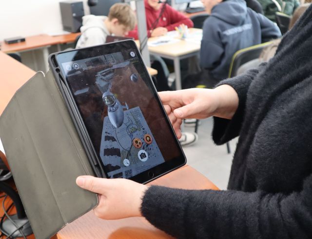 Na zdjęciu znajduje się tablet, za pomocą którego kobieta ożywia ilustrację. W tle znajduje się młodzież przy stolikach.