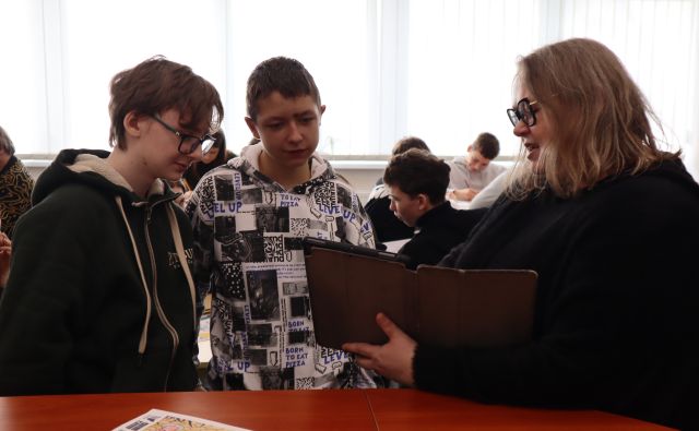 Na zdjęciu znajduje sie kobieta trzymająca tablet i pokazuje coś dwóm chłopakom, którzy stoją obok niej. W tle znajduje się reszta uczestników warsztatów oraz okna w tle.