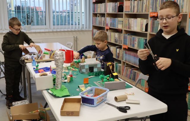 Na zdjęciu znajduje się trójka chłopców przy złączonych stołach, jeden z nich siedzi i wspólnie tworzą kolejne elementy do makiety miasta, znajdującej się na stole wraz z nożyczkami, kartonikami. W tle znajdują się okna i półki z książkami.