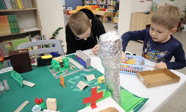 Na zdjęciu znajduje się dwójka chłopców przy stole, jeden z nich się nachyla przy stole i tworzy mały element do makiety miasta, która stoi przed chłopcami, na makiecie znajdują się drzewa, wieża ciśnień, szpital. W tle znajdują się półki z książkami.