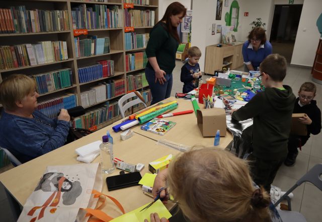 Na zdjęciu znajduje się trójka chłopców oraz cztery kobiety przy stole i wspólnie tworzą dodatkowe elementy do makiety z kolorowych kartek, kartoników i bibuły znajdujących się na stole. Dwie kobiety oraz jeden jeden chłopiec stoją przy stole. W tle znajdują się półki z książkami.