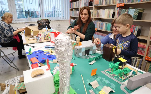 Na zdjęciu znajdują się trzy kobiety oraz jeden chłopiec przy złączonych stolikach, na których leży makieta miasta z budynkami, roślinnością i wieżą ciśnień. W tle znajdują się okna i półki z książkami.