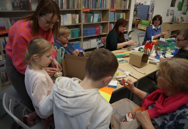 Na zdjęciu znajdują się dzieci wraz z dorosłymi przy złączonych stołach i wspólnie tworzą budynki do makiety miasta, która znajdują się na stole wraz z bibułą, kredkami, kolorowymi kartkami i kartonikami. W tle znajdują się półki z książkami.