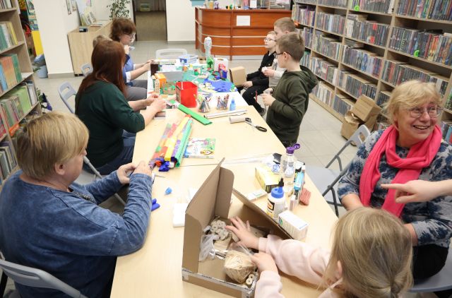 Na zdjęciu znajdują się cztery kobiety oraz piątka dzieci przy złączonych stołach, na których leżą kartoniki, kolorowa bibuła, kleje oraz makieta miasta na końcu stołu, do której tworzą elementy. Po lewej i prawej stronie znajdują się półki z książkami.