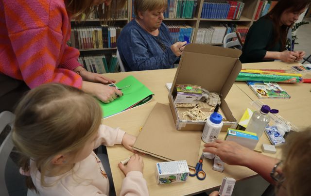Na zdjęciu znajdują się cztery kobiety przy stole, jedna z nich nachyla się nad zieloną kartką, a obok z niej siedzi dziewczynka. Na złączonych stolikach znajdują się kartoniki, kolorowa bibuła, kleje. W tle znajdują się półki z książkami.