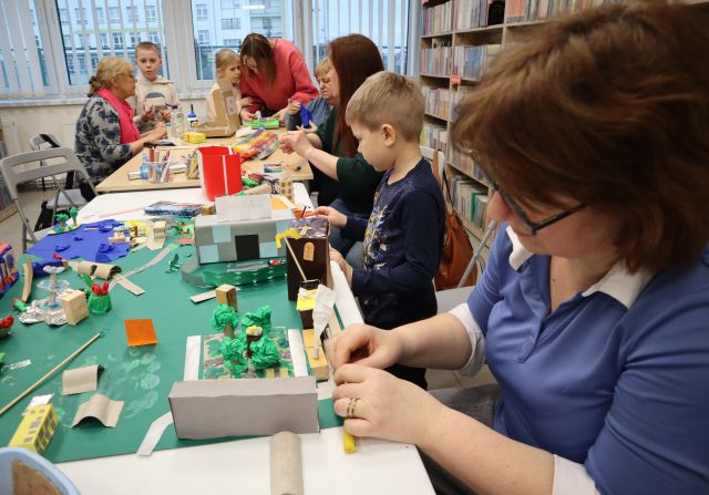Na zdjęciu znajduje się pięć kobiet oraz trójka dzieci przy złączonych stołąch. Jedna kobieta siedzi przed makietą i dokleja do niej kolejne elementy, a reszta tworzy elementy do makiety z kartoników, bibuły i kredek. W tle znajdują się okna, a po prawej stronie półki z ksiażkami.