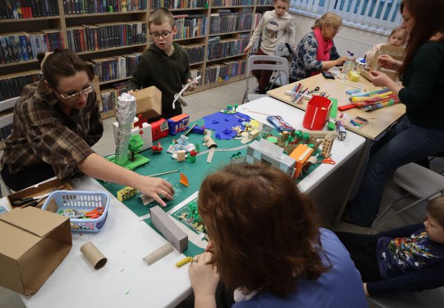 Na zdjęciu znajdują się kobiety wraz z dziećmi przy złączonych stołach i wspólnie tworzą dodatkowe elementy do makiety miasta z bibuły, kartoników. Przy makiecie, na której znajdują się budynki, jezioro, wieża ciśnień siedzą dwie kobiety i doklejają kolejne elementy. W tle znajdują się półki z książkami.