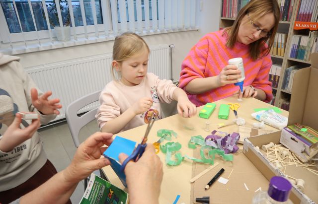 Na zdjęciu znajduje się kobieta wraz z dziewczynką przy stole i z kartoników i kolorowych kartek robią budynki. Obok znajdują się ręce kobiety, która wycina kwadrat z niebieskiej kartki, a obok niej stoi chłopiec. Na stole znajdują się wycinki z bibuły, kartoniki oraz klej. W tle znajdują się okna i półki z książkami.