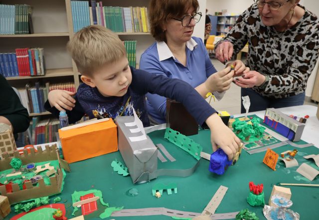 Na zdjęciu przy złączonych stołach znajduje się chłopiec doklejający niebieski budynek do makiety, obok niego znajdują się dwie kobiety i wspólnie tworzą kolejny element makiety. Makieta miasta zawiera budynki, drogi, roślinność. W tle znajdują się półki z książkami.