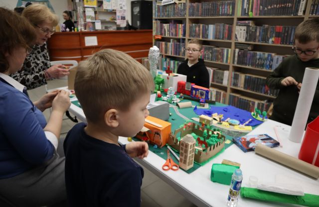 a zdjęciu znajdują się dwie kobiety oraz trójka chłopców po dwóch stronach złączonych stołów, na któruch leży makieta miasta wraz z budynkami, roślinnością i jeziorem. Uczestnicy warsztatów robią dodatkowe elementy z bibuły. W tle znajdują się półki z książkami.
