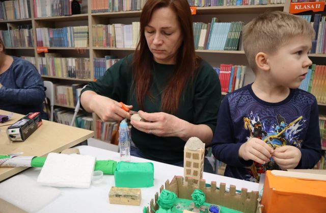 Na zdjęciu znajduje się kobieta wraz z chłopcem przy stole, na którym leży makieta miasta. Kobieta przecina mały kawałek rolki po papierze, a chłopiec jest odwrócony w prawą stronę. Na makiecie znajdują się budynki i roślinność. W tle znajdują się półki z książkami.