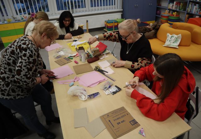 Na zdjęciu znajdują się dwie dziewczyny oraz trzy kobiety przy stole, które wycinają z materiałów znajdujących się na stole wraz z kartonikami oraz nitkami. W tle znajdują się okna, po prawej stronie okrągła kanapa.