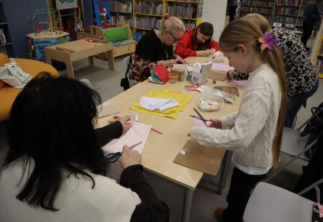 Na zdjęciu znajdują się dwie dziewczynki oraz trzy kobiety, które wycinają wzory z materiałów znajdujących się na stole wraz z nitkami i nożyczkami. W tle znajdują się półki z książkami. 