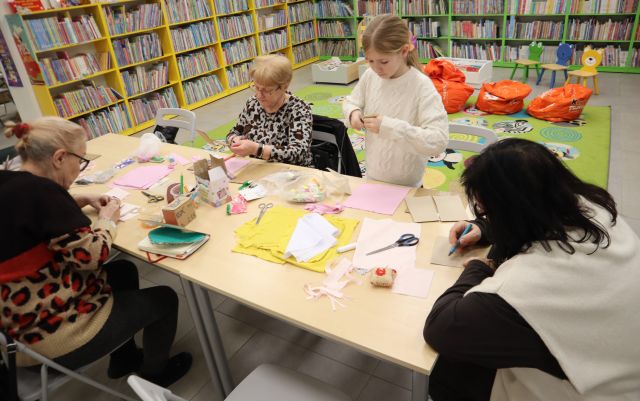 Na zdjęciu znajdują się trzy kobiety oraz jedna stojąca dziewczynka. Uczestniczki zajęć wycinają z materiałów wzór do szycia. Na stole znajdują się żółte i różowe materiały, nożyczki oraz kreda. W tle znajduje się dywan z pufami i półki z książkami.