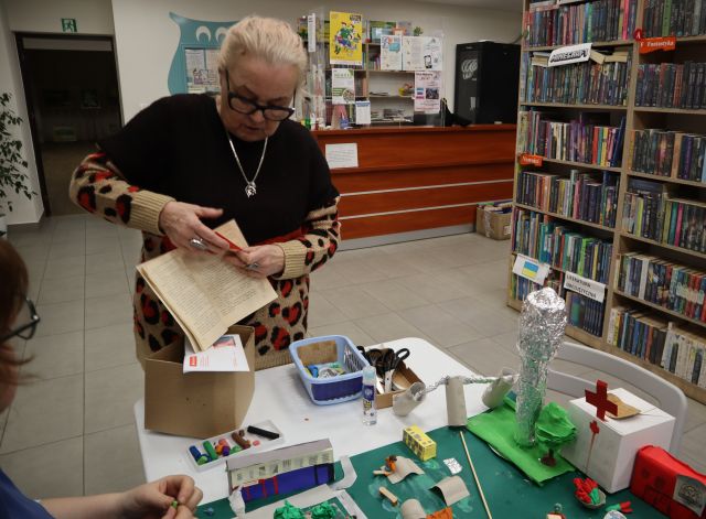 Na zdjęciu znajduje się kobieta, która trzyma w ręce otwartą starą książkę. Na stole, przy którym stoi kobieta znajduje się karton, nożyczki, plastelina oraz makieta miasta z budynkami, wieżą ciśnień i roślinnością. Przy stole po lewej stronie siedzi kobieta. Po prawej stronie znajdują się półki z książkami, a w tle biurko.
