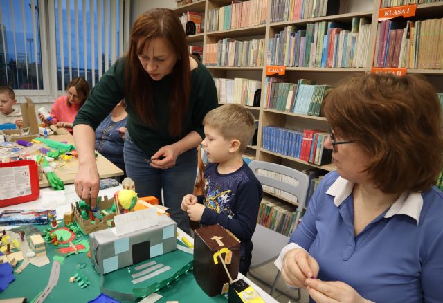 Na zdjęciu w centrum znajdują się dwie kobiety i chłopiec między nimi, znajdują się przy stole jedna kobieta stoi i wspólnie tworzą elementy do makiety miasta. Na złączonych stołach w tle znajduje się bibuła, pudełka oraz trójka uczestników. W tle znajdują się okna, a po prawej stronie półki z książkami.