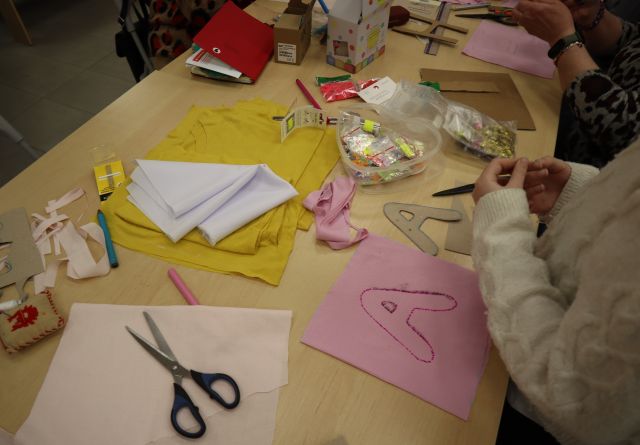 Na zdjęciu znajdują się złączone stoły z materiałami, nożyczkami i koralikami, na brzegu stołu leży materiał z wyszytą literką A, a przy stole stoi dziewczynka. Zdjęcie zostało wykonane od góry.