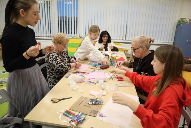 Na zdjęciu znajdują się dwie dziewczynki, jedna z nich stoi i sięga materiał z końca stołu, przy stole siedzą również trzy kobiety. a lewej stronie stoi kobieta z nitką w ręce. Na złączonych stolikach leżą materiały, kredy i pisaki. W tle znajdują się okna.