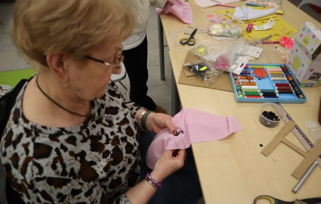 Na zdjęciu znajduje się kobieta, która przyszywa koraliki do różowego materiału, na złączonych stolikach przed nią leżą nitki, wycięte H z kartonu oraz nożyczki i inne materiały. Zdjęcie zostało wykonane od boku.