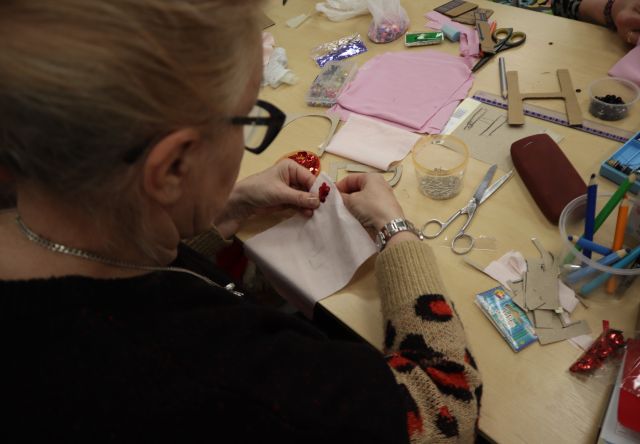 Na zdjęciu znajduje się kobieta przy stole, która przyszywa koraliki do różowego materiału, na stole przed kobietą leżą różowe materiały, nożyczki, kredki i koraliki. Zdjęcie zostało wykonane od tyłu.