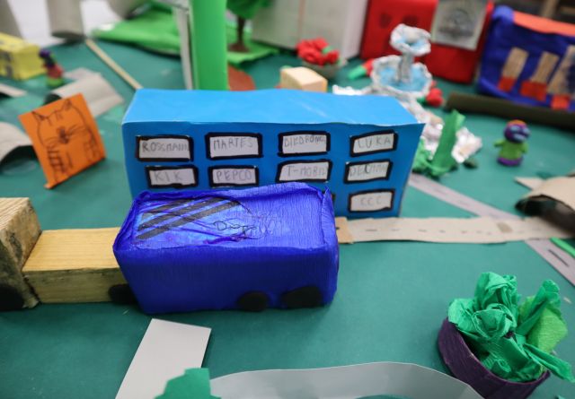 Na zdjęciu znajduje się część makiety przedstawiająca niebieski budynek, drogi, niebieski samochód, roślinność.