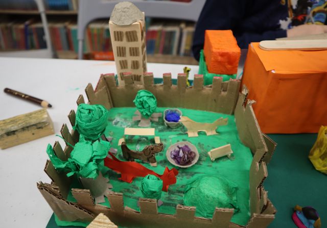 Na zdjęciu znajduje się kawałek makiety przedstawiający zagrodę z pieskami, drzewami. Obok stoją budynki.