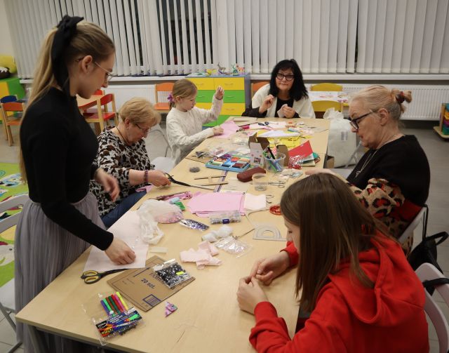 Na zdjęciu znajdują się dwie dziewczynki siedzące przy stole wraz z trzema kobieta. Jedna kobieta stoi przy stole, na których leżą różowe materiały, kredki, nitki. W tle znajdują się okna.