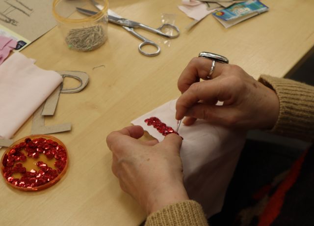 Na zdjęciu znajdują się dłonie kobiety, któa przyszywa czerwone koraliki do różowego materiału. Na stole przed kobietą leżą nożyczki, koraliki oraz resztki materiału.